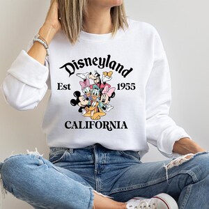 Puede incluir: Sudadera blanca con un dise&ntilde;o colorido de personajes de Disney, incluyendo Mickey y Minnie Mouse, Donald Duck y Goofy. El texto "Disneyland Est 1955 CALIFORNIA" est&aacute; impreso en negro.