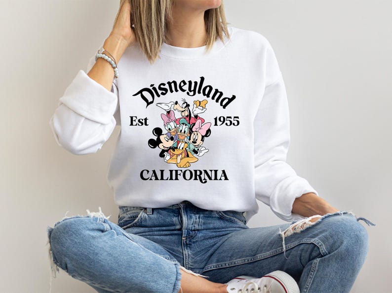Puede incluir: Sudadera blanca con un dise&ntilde;o colorido de personajes de Disney, incluyendo Mickey y Minnie Mouse, Donald Duck y Goofy. El texto "Disneyland Est 1955 CALIFORNIA" est&aacute; impreso en negro.