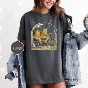 Puede incluir: Camiseta gris oscuro con una ilustraci&oacute;n caprichosa de dos ranas en bicicleta. La obra de arte est&aacute; enmarcada por un arco con detalles florales. La camiseta tiene un corte holgado y est&aacute; hecha de un material suave.