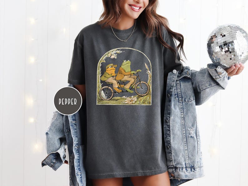 Puede incluir: Camiseta gris oscuro con una ilustraci&oacute;n caprichosa de dos ranas en bicicleta. La obra de arte est&aacute; enmarcada por un arco con detalles florales. La camiseta tiene un corte holgado y est&aacute; hecha de un material suave.