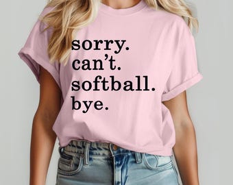 May include: A pale pink t-shirt with the words "sorry. can't. softball. bye." printed in black. The shirt has short sleeves and a round neck.