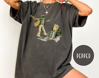 May include: Dark grey t-shirt featuring a whimsical illustration of two frogs. One frog is standing with arms outstretched, while the other sits. The shirt has a relaxed fit and the word "PEPPER" is printed on a dark grey circle.