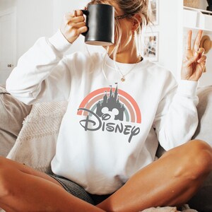 May include: White sweatshirt featuring a colorful rainbow design with a castle silhouette, Mickey Mouse ears, and the word "Disney" in gray. The sweatshirt has long sleeves and a crew neck. A black mug is held up to the face.