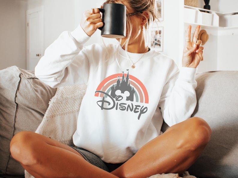 May include: White sweatshirt featuring a colorful rainbow design with a castle silhouette, Mickey Mouse ears, and the word "Disney" in gray. The sweatshirt has long sleeves and a crew neck. A black mug is held up to the face.