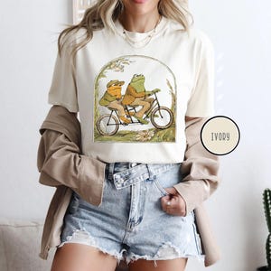 Puede incluir: Camiseta de color marfil con una ilustraci&oacute;n caprichosa de dos ranas montando en bicicleta t&aacute;ndem. Las ranas visten ropa de &eacute;poca, sobre un fondo de vegetaci&oacute;n y flores. La camiseta tiene cuello redondo y mangas cortas.
