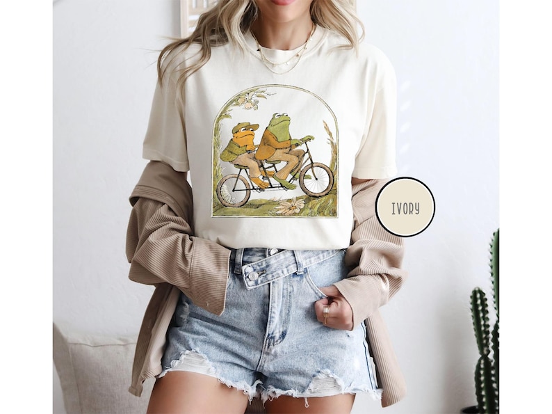 Puede incluir: Camiseta de color marfil con una ilustraci&oacute;n caprichosa de dos ranas montando en bicicleta t&aacute;ndem. Las ranas visten ropa de &eacute;poca, sobre un fondo de vegetaci&oacute;n y flores. La camiseta tiene cuello redondo y mangas cortas.