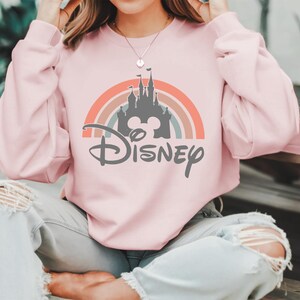 May include: A light pink sweatshirt featuring a rainbow design with a gray castle silhouette and the word "Disney" in a stylized font. The sweatshirt has long sleeves.