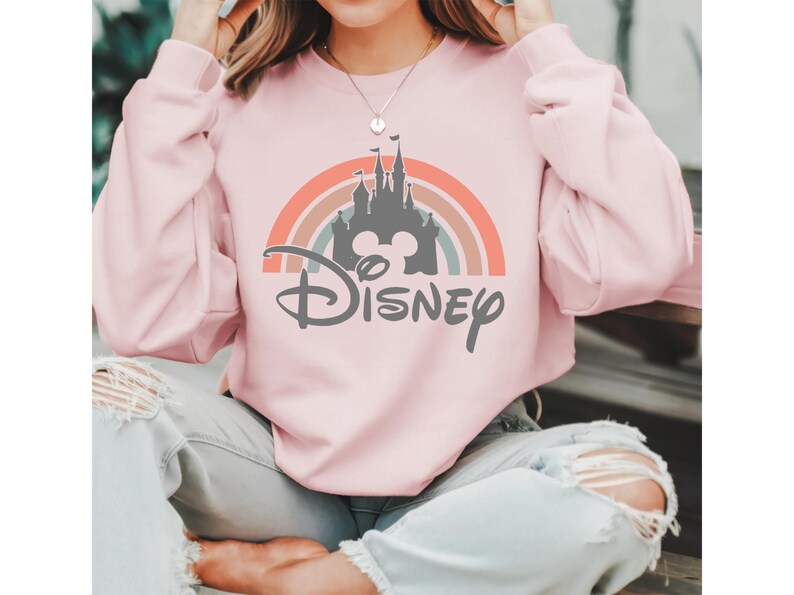 May include: A light pink sweatshirt featuring a rainbow design with a gray castle silhouette and the word "Disney" in a stylized font. The sweatshirt has long sleeves.