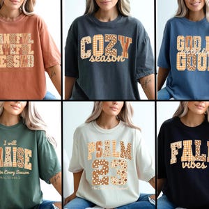May include: Collection of six t-shirts in various colors, each featuring a different text design. The designs include phrases like "Thankful, Grateful, Blessed," "Cozy Season," and "God is all the time good." The text is in a patchwork style.