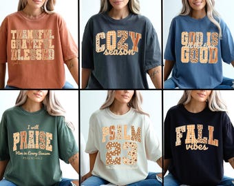 May include: Collection of six t-shirts in various colors, each featuring a different text design. The designs include phrases like "Thankful, Grateful, Blessed," "Cozy Season," and "God is all the time good." The text is in a patchwork style.