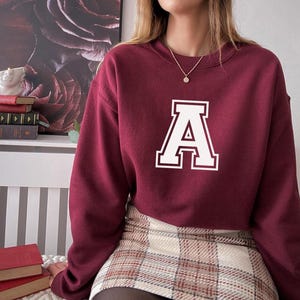 May include: Burgundy crewneck sweatshirt with a large white letter "A" on the front. The sweatshirt is paired with a plaid mini skirt. A gold necklace is visible.