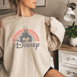 May include: Beige sweatshirt featuring a colorful graphic of a castle, rainbow, and the word "Disney" in a stylized font. The design incorporates pastel colors. The sweatshirt is a casual, long-sleeved garment.