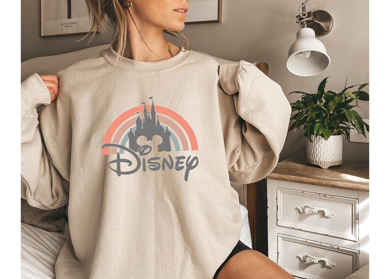 May include: Beige sweatshirt featuring a colorful graphic of a castle, rainbow, and the word "Disney" in a stylized font. The design incorporates pastel colors. The sweatshirt is a casual, long-sleeved garment.