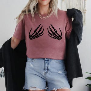 May include: A mauve-colored t-shirt featuring a black skeleton hand design on the front. The graphic depicts two hands in a gesture. The model is wearing denim shorts and a black jacket.