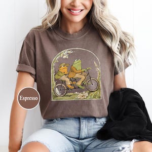 Puede incluir: Camiseta color espresso con una ilustraci&oacute;n caprichosa de dos ranas montando en bicicleta. El dise&ntilde;o est&aacute; enmarcado por un arco con detalles florales. La camiseta la lleva una persona.