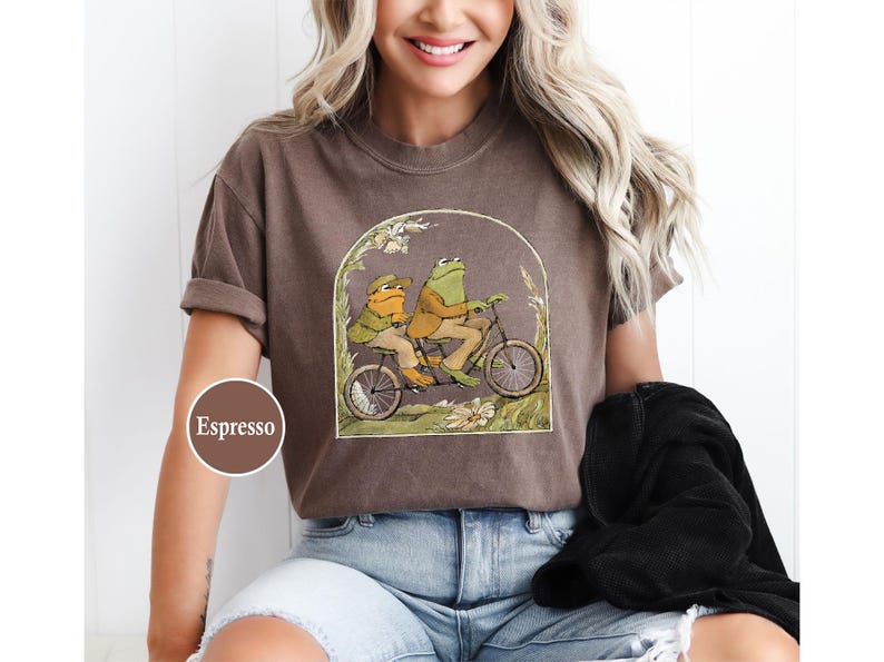 Puede incluir: Camiseta color espresso con una ilustraci&oacute;n caprichosa de dos ranas montando en bicicleta. El dise&ntilde;o est&aacute; enmarcado por un arco con detalles florales. La camiseta la lleva una persona.