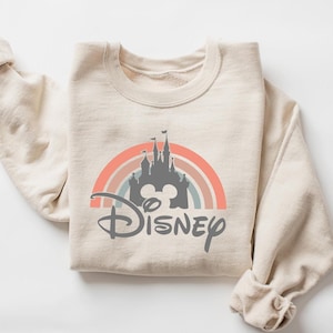 May include: Beige sweatshirt featuring a colorful rainbow design with a gray castle silhouette and the word "Disney" in a script font. The sweatshirt is folded, with the sleeves visible.