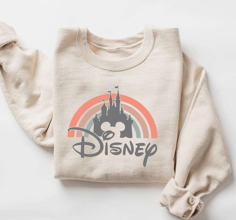 May include: Beige sweatshirt featuring a colorful rainbow design with a gray castle silhouette and the word "Disney" in a script font. The sweatshirt is folded, with the sleeves visible.