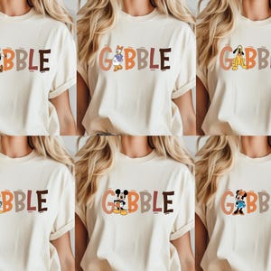 May include: Cream-colored t-shirts with the word "GOBBLE" in colorful letters. Each shirt features a different Disney character: Goofy, Daisy Duck, Pluto, Donald Duck, Mickey Mouse, and Minnie Mouse. The shirts are short-sleeved.