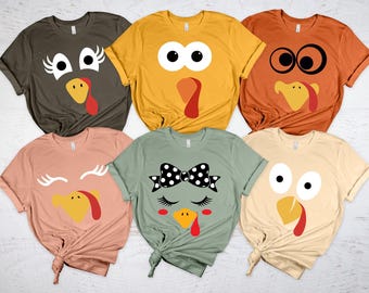 May include: Six colorful t-shirts with turkey face designs. The shirts are in various colors, including olive green, mustard yellow, and peach. Each shirt features a different turkey face design, perfect for Thanksgiving or fall.