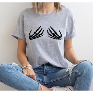May include: A light gray t-shirt featuring a black graphic of two skeleton hands. The hands are positioned as if cupping the chest. The shirt has short sleeves and a relaxed fit, suitable for casual wear.