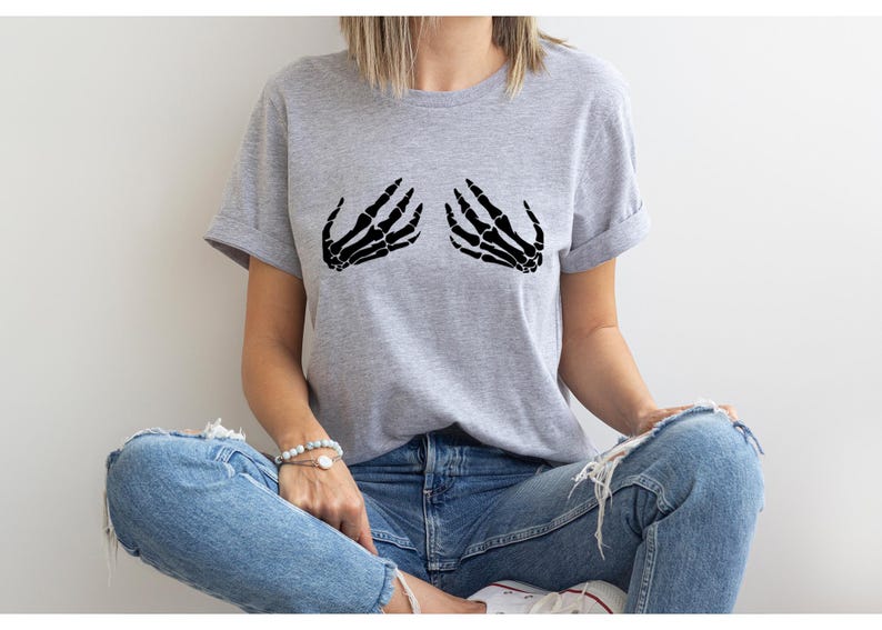 May include: A light gray t-shirt featuring a black graphic of two skeleton hands. The hands are positioned as if cupping the chest. The shirt has short sleeves and a relaxed fit, suitable for casual wear.