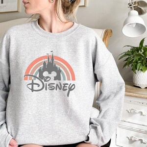 May include: A light gray sweatshirt featuring a colorful rainbow design with a castle silhouette and the word "Disney" in a script font. The design incorporates shades of orange, pink, and blue.