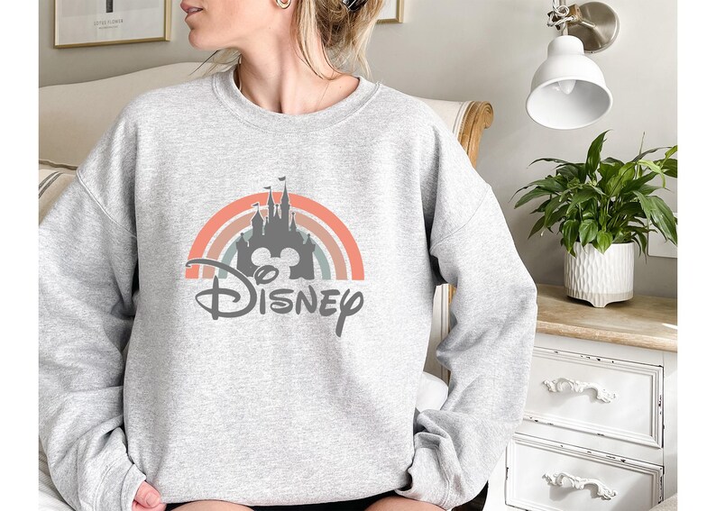 May include: A light gray sweatshirt featuring a colorful rainbow design with a castle silhouette and the word "Disney" in a script font. The design incorporates shades of orange, pink, and blue.