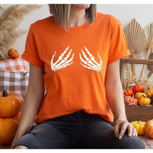 May include: Orange t-shirt with rolled sleeves featuring a white skeleton hand design. The shirt is part of a Halloween or fall-themed collection. The model is wearing dark jeans.