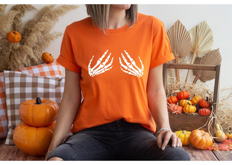 May include: Orange t-shirt with rolled sleeves featuring a white skeleton hand design. The shirt is part of a Halloween or fall-themed collection. The model is wearing dark jeans.