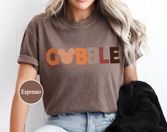 May include: Espresso-colored t-shirt with the word "GOBBLE" in orange, tan, and brown letters. The letter "O" is designed with a Mickey Mouse head shape. The shirt is a casual style.
