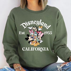 Puede incluir: Una sudadera verde oliva con un gr&aacute;fico de personajes de Disney, incluyendo Mickey y Minnie Mouse, Donald Duck y Goofy. El texto "Disneyland Est 1955 CALIFORNIA" est&aacute; impreso en blanco sobre los personajes.