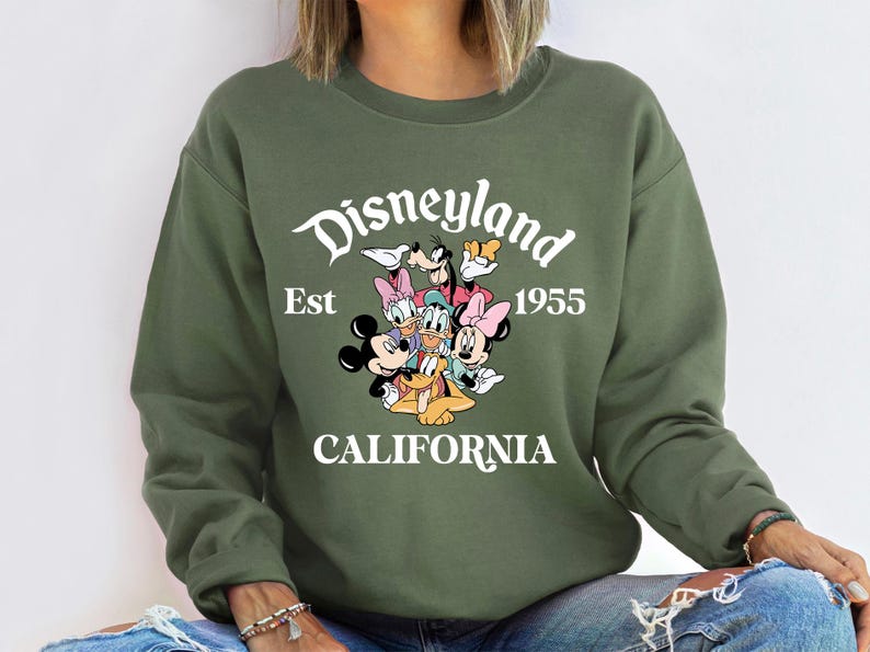 Puede incluir: Una sudadera verde oliva con un gr&aacute;fico de personajes de Disney, incluyendo Mickey y Minnie Mouse, Donald Duck y Goofy. El texto "Disneyland Est 1955 CALIFORNIA" est&aacute; impreso en blanco sobre los personajes.