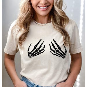 May include: Beige t-shirt featuring a black skeleton hand graphic on the front. The graphic depicts two hands in a gesture. The shirt is made of a soft material and has short sleeves. The overall style is casual.