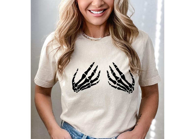 May include: Beige t-shirt featuring a black skeleton hand graphic on the front. The graphic depicts two hands in a gesture. The shirt is made of a soft material and has short sleeves. The overall style is casual.