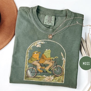 Puede incluir: Una camiseta verde musgo Comfort Colors con una ilustraci&oacute;n de dos ranas en bicicleta t&aacute;ndem. La camiseta tiene cuello redondo y mangas cortas. Tambi&eacute;n se ven un collar dorado y un sombrero marr&oacute;n.