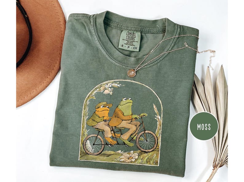 Puede incluir: Una camiseta verde musgo Comfort Colors con una ilustraci&oacute;n de dos ranas en bicicleta t&aacute;ndem. La camiseta tiene cuello redondo y mangas cortas. Tambi&eacute;n se ven un collar dorado y un sombrero marr&oacute;n.