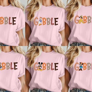 May include: Pink t-shirts with the word "GOBBLE" in colorful letters and various cartoon characters. The characters include Goofy, Daisy Duck, Pluto, Donald Duck, Mickey Mouse, and Minnie Mouse. The shirts are casual wear.