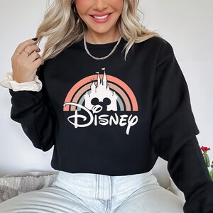 May include: Black crewneck sweatshirt featuring a colorful graphic of a castle, a rainbow, and the word "Disney" in white script. The design includes the iconic Mickey Mouse silhouette. A casual, comfortable top.