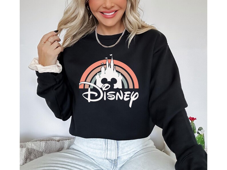 May include: Black crewneck sweatshirt featuring a colorful graphic of a castle, a rainbow, and the word "Disney" in white script. The design includes the iconic Mickey Mouse silhouette. A casual, comfortable top.