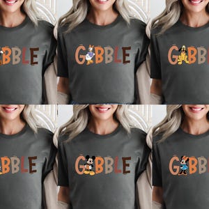 May include: A dark gray t-shirt with the word "GOBBLE" in orange and brown letters. Each letter features a different cartoon character, including Goofy, Donald Duck, Pluto, Daisy Duck, Mickey Mouse, and Minnie Mouse.