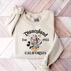 Puede incluir: Sudadera color crema con la palabra "Disneyland" en fuente cursiva, con "Est 1955" y "CALIFORNIA" debajo. Un gr&aacute;fico de personajes de Disney est&aacute; en el centro. La sudadera tiene mangas largas y cuello redondo.