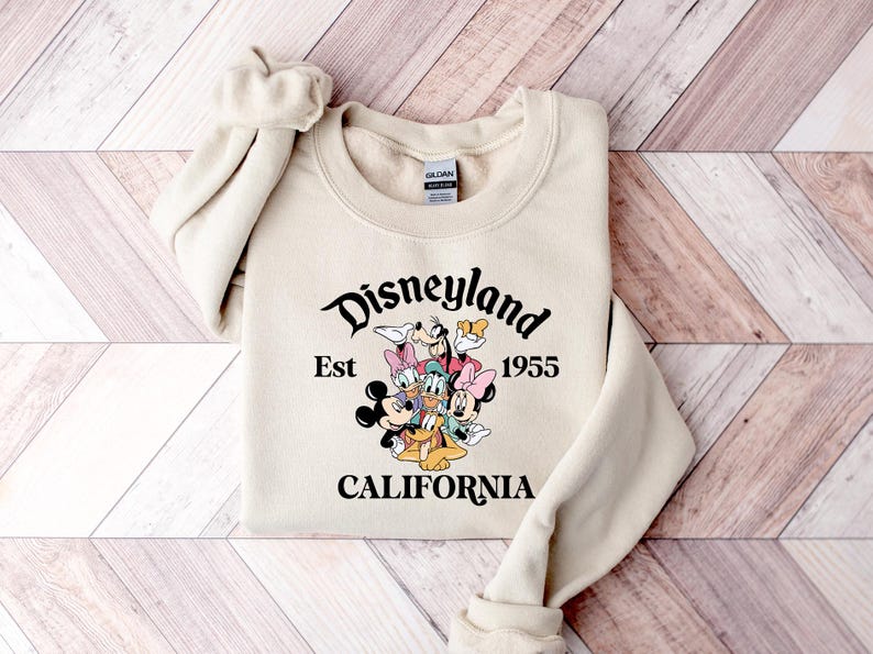 Puede incluir: Sudadera color crema con la palabra "Disneyland" en fuente cursiva, con "Est 1955" y "CALIFORNIA" debajo. Un gr&aacute;fico de personajes de Disney est&aacute; en el centro. La sudadera tiene mangas largas y cuello redondo.