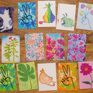May include: Set of 12 illustrated greeting cards featuring various designs, including cats, flowers, plants, and peace signs. The cards are printed on white cardstock and have a blank inside for writing.