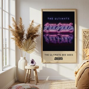Puede incluir: Un póster enmarcado con el texto "THE ULTIMATE BEE GEES" en una fuente de estilo neón. El póster está en un marco de madera de color claro y cuelga de una pared sobre una cama. La habitación tiene una paleta de colores neutros.