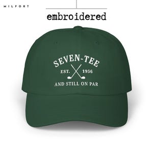 May include: A dark green baseball cap featuring white embroidery on the front. The text reads SEVEN-TEE EST. 1956 AND STILL ON PAR, accompanied by a graphic of two crossed golf clubs. The word embroidered is printed above the cap.