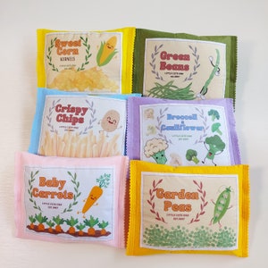 May include: Set of ten felt food pouches with colorful illustrations of various vegetables. Each pouch features a different vegetable, including sweet corn, green beans, crispy chips, broccoli and cauliflower, baby carrots, and garden peas. The pouches are labeled with the name of the vegetable and the phrase "Little Cute One".