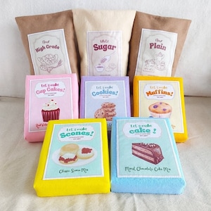 Puede incluir: Un conjunto de bolsas de comida de fieltro para juegos de simulación. Las bolsas son de color marrón, beige y amarillo y presentan imágenes de cupcakes, galletas, muffins, scones y pastel. Las bolsas están etiquetadas con el texto "Harina de alta calidad", "Azúcar blanco", "Harina simple", "¡Hagamos cupcakes!", "¡Hagamos galletas!", "¡Hagamos muffins!", "¡Hagamos scones!" y "¡Hagamos un pastel!"