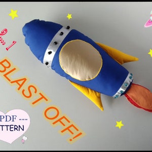 May include: A blue and yellow rocket-shaped plush toy with a gold star on the front. The toy has a red and orange flame tail. The text "BLAST OFF!" is written in orange on a yellow background. The text "--PDF-- PATTERN" is written in black on a pink heart.