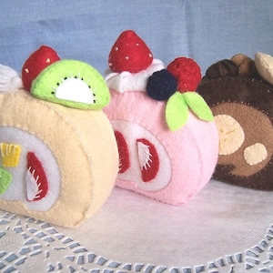 Puede incluir: Tres alimentos de fieltro con forma de pastel de fresa, rollo de kiwi y rollo de chocolate. El pastel de fresa es rosa con glaseado blanco y una hoja verde. El rollo de kiwi es amarillo con una rodaja de kiwi verde en la parte superior. El rollo de chocolate es marrón con un remolino blanco.